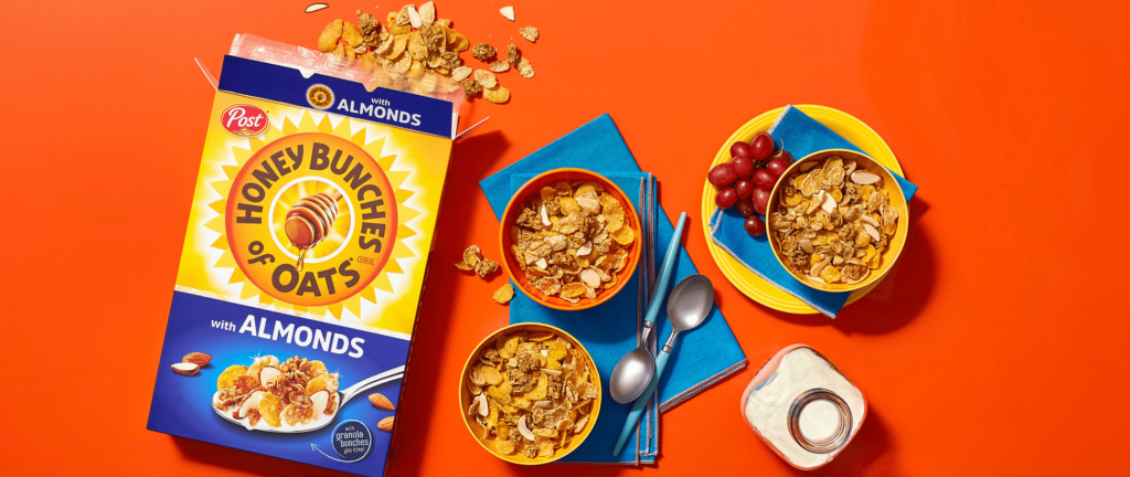 Honey Bunches of Oats with Almonds cereal in a box and bowls next to a small bottle of milk