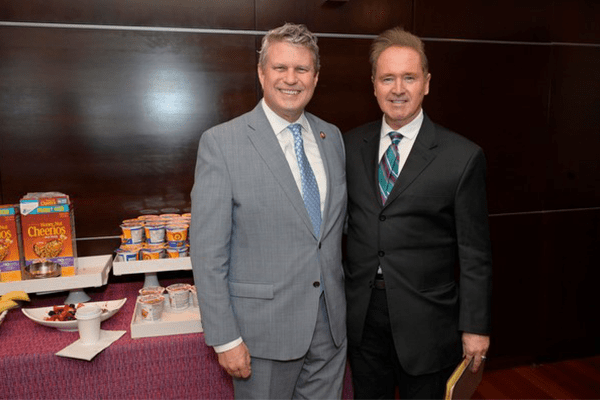 Two men in suits stand by a table where Honey Bunches of Oats can be seen.