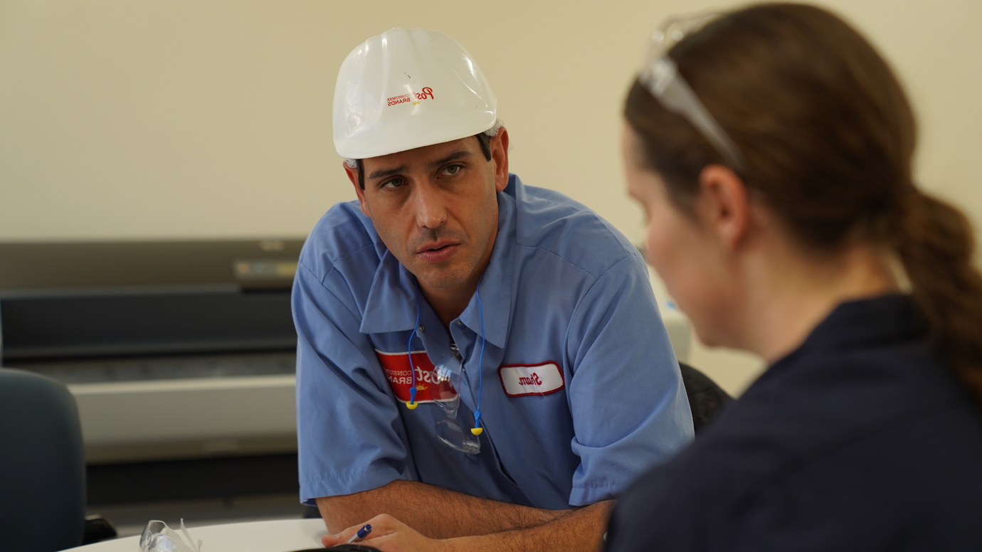 A Post employee in a hardhat talks to a woman in a black shirt