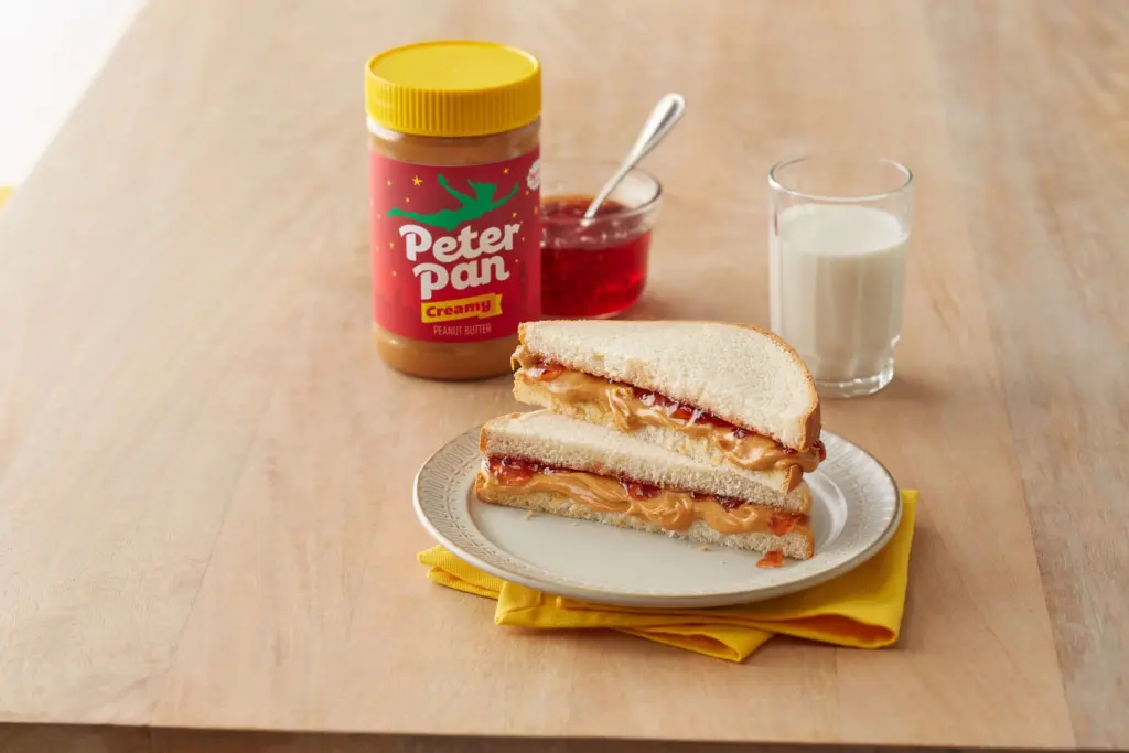 Creamy Peter Pan Peanut Butter next to a glass of milk and peanut butter and jelly sandwich