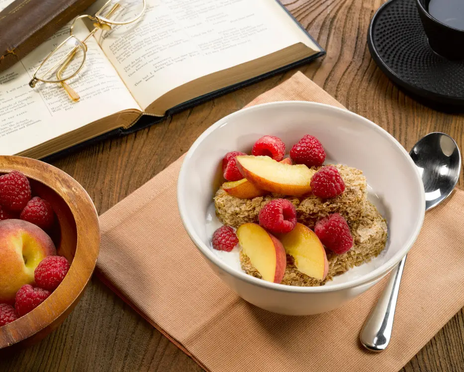 a bowl of Weetabix cereal with peaches and raspberries next to an open book